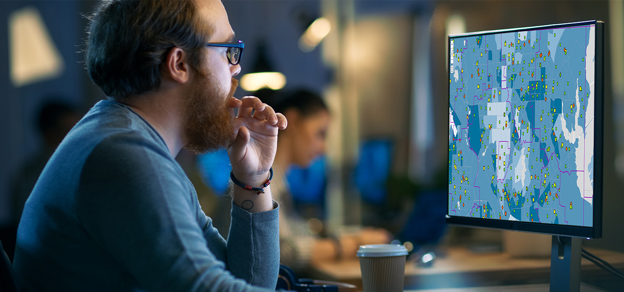 Człowiek w niebieskiej koszulce z długim rękawem siedzący przy biurku we wspólnej przestrzeni biurowej i oglądający cyfrową mapę na monitorze