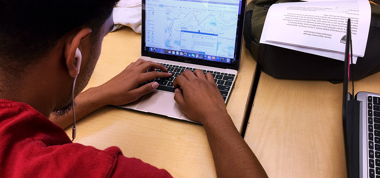 Студент в наушниках и красной футболке сидит за классным столом и просматривает цифровую карту на маленьком ноутбуке