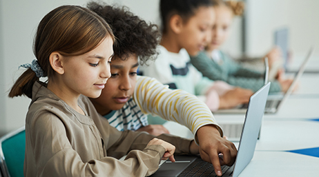 Four children sitting in a row at a long desk sharing several laptops in a brightly lit modern classroom