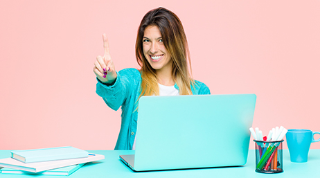 A smiling person wearing a sky-blue sweater holding a finger up in a “hold on” gesture while working at a blue desk with blue accessories and a blue laptop with a pale pink background