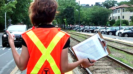 A person holding a clipboard and wearing an orange and yellow safety vest at a street roadside making a thumbs-up gesture at an approaching car