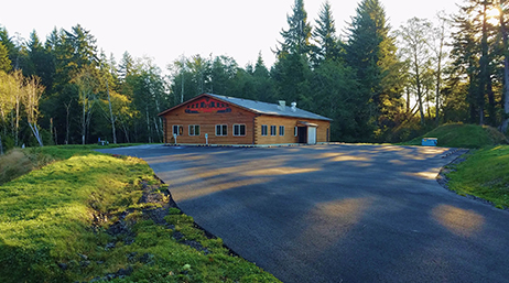 A building in the middle of a paved lot surrounded by pine trees and greenery