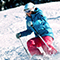 Skier in a blue jacket, red pants, and helmet and goggles leans into the slope and kicks up snow during a maneuver