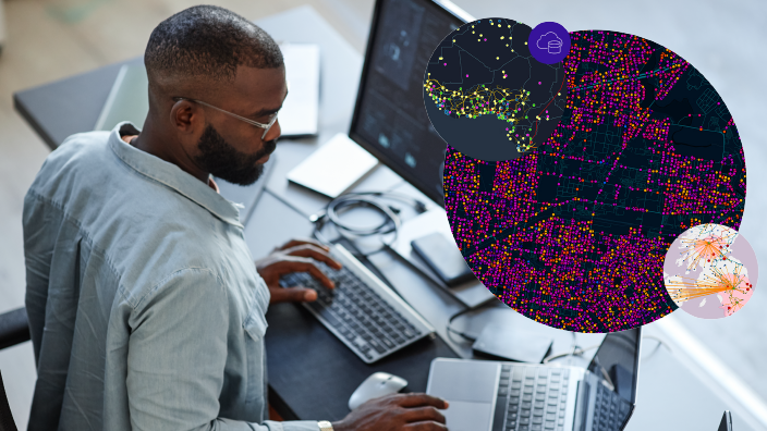 A person in a light gray collared shirt seated at a sleek modern desk with several monitors displaying colorful map data