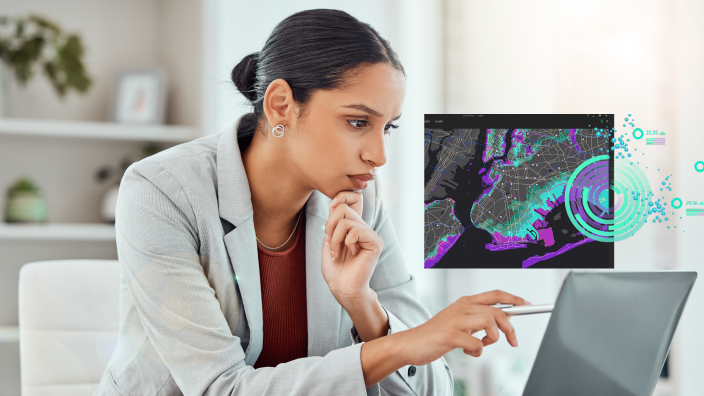 A person wearing a pale gray suit and red blouse sitting in a bright modern office using a laptop, overlaid with a small colorful map