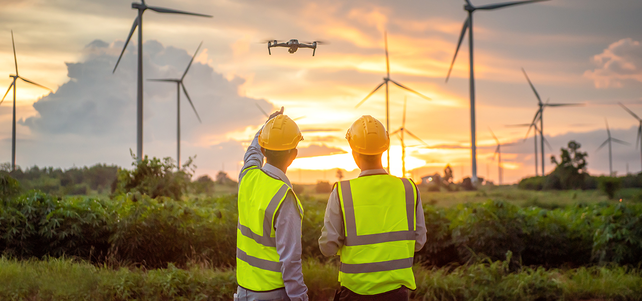 The backs of two people wearing safety vests and hard hats looking up at a drone flying near a handful of wind turbines