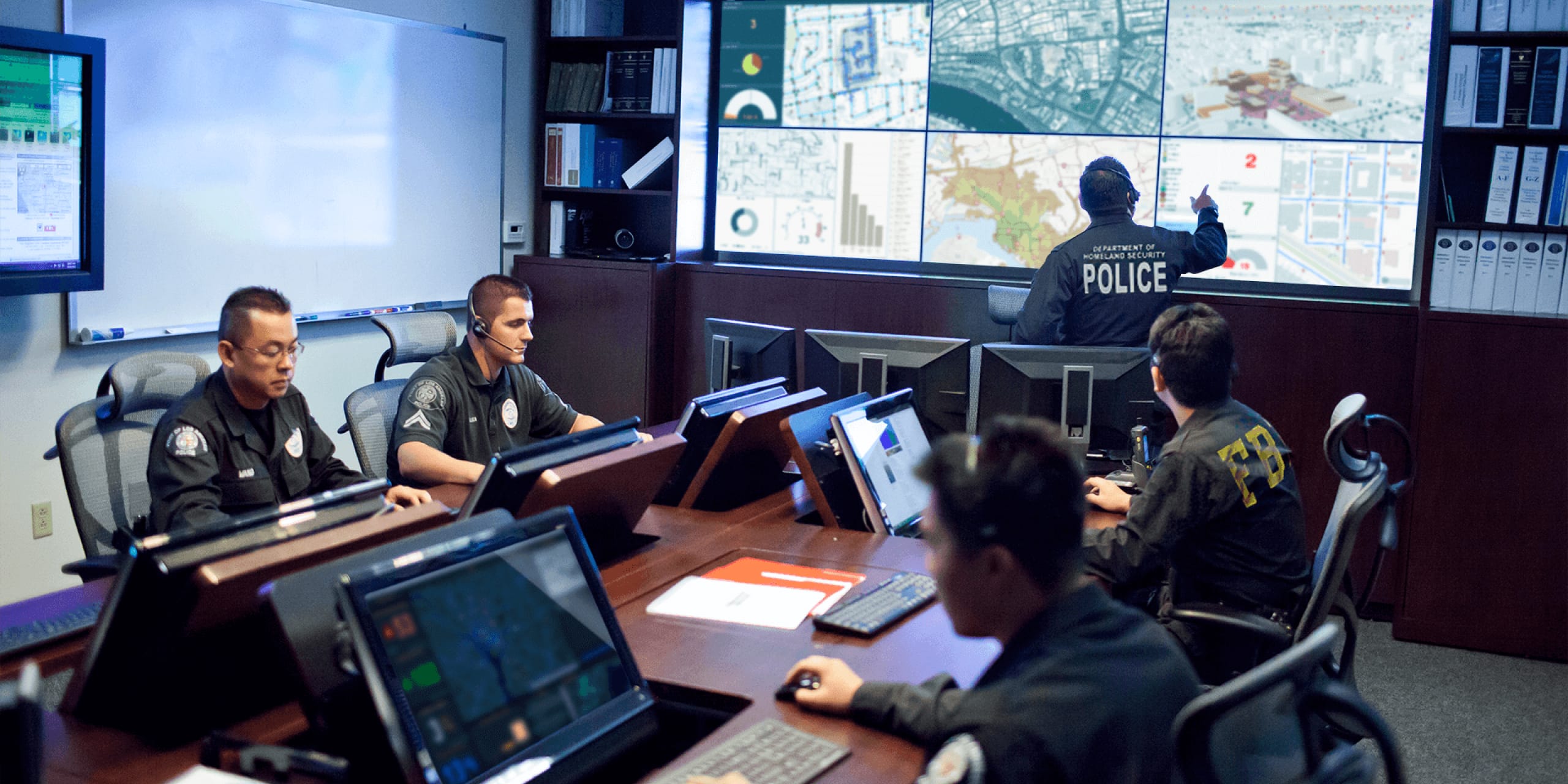 Law enforcement officers sitting at a conference table using computers under a large display of different digital maps