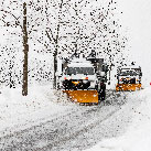 Two yellow snowplows pushing snow off a city street during heavy snowfall in a landscape completely covered in deep snow