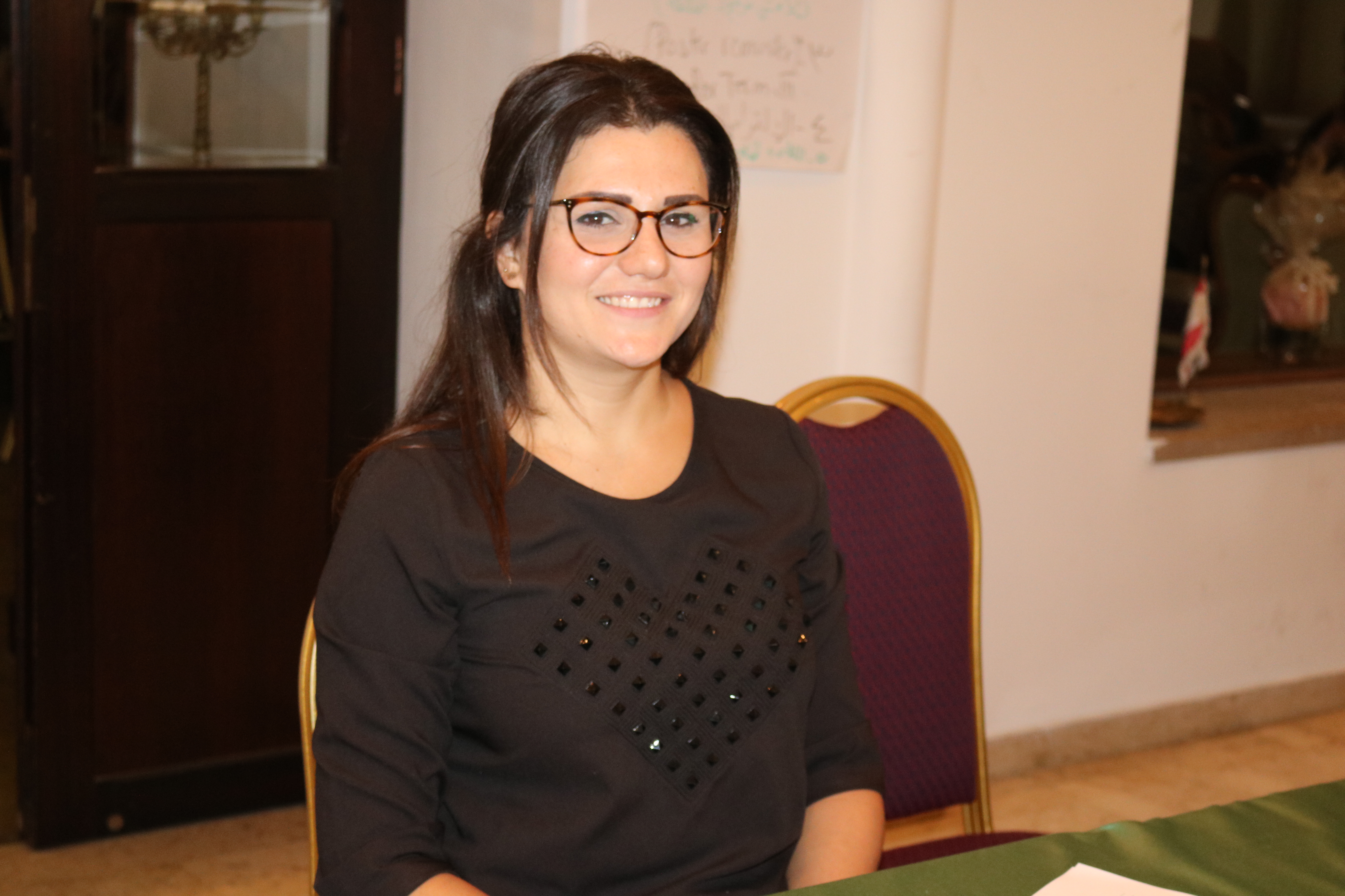 Mayssam Imad, smiling, looks directly at the camera, has long black hair, is wearing brown round glasses and a black shirt