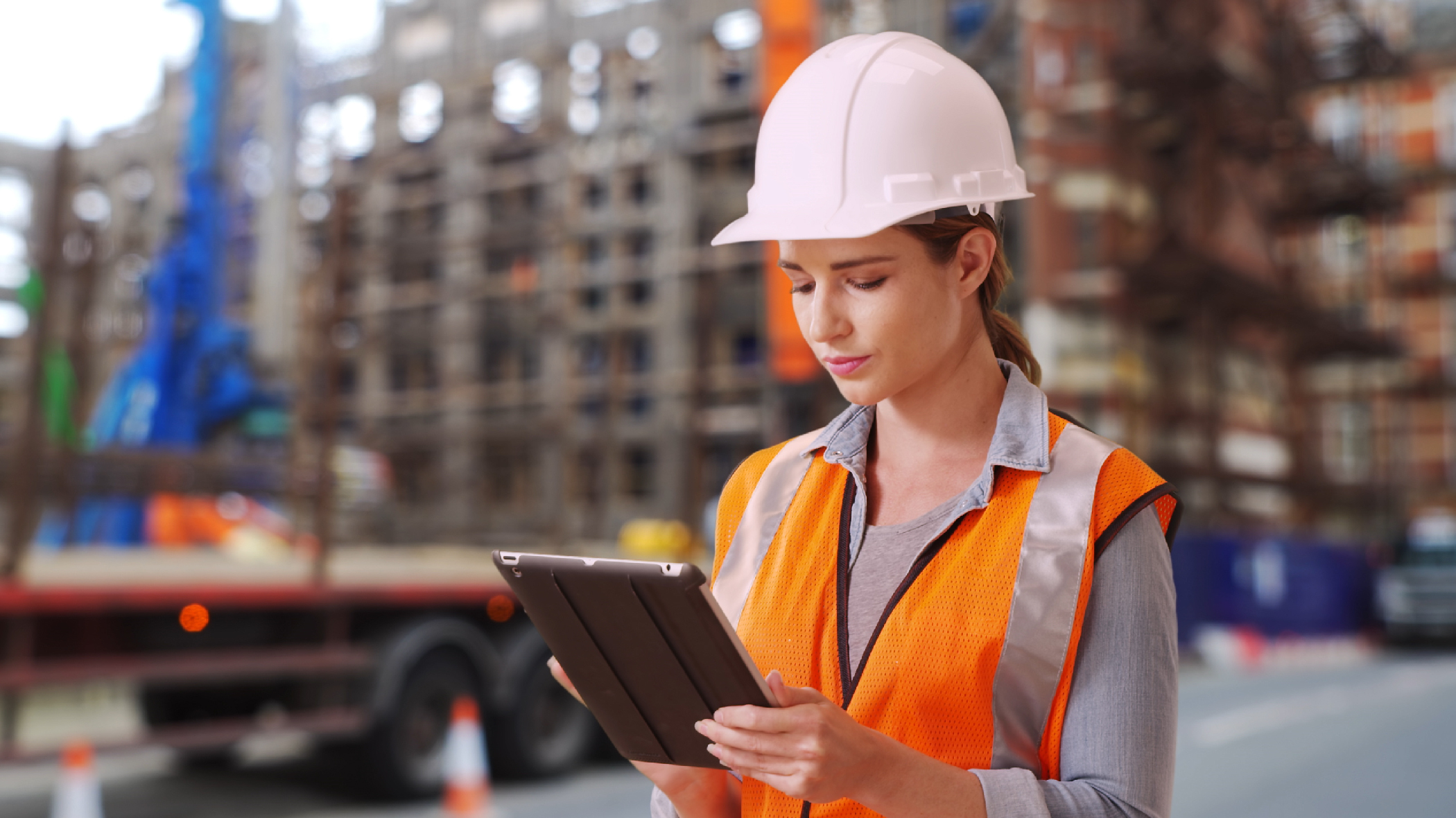 Woman wearing a hard hat and safetly vest holds a tablet device in her hand at an outdoor construction site.