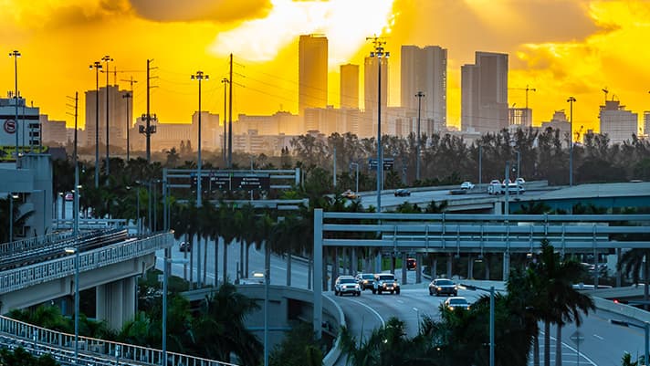 Miami freeway lined by palm trees with the downtown skyline visible against a vibrant orange sky at sunset