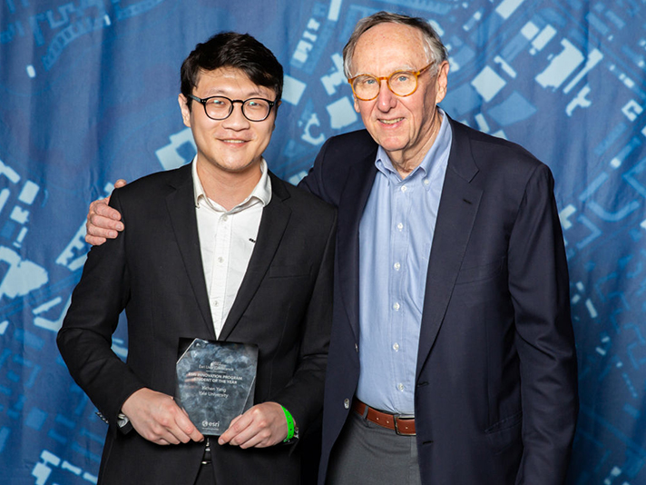 Yale student Yichen Yang holding an award and standing with Esri president Jack Dangermond in front of a blue backdrop