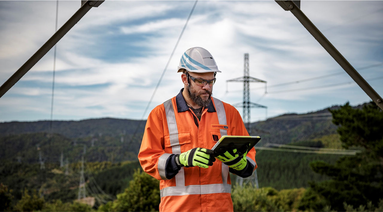 Photo of a man with glasses in a hard hat with orange jacket and rubber gloves on a tablet 