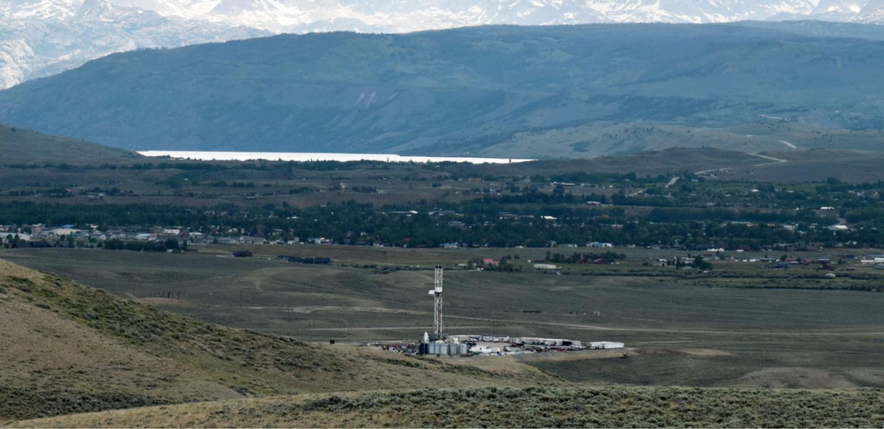 A well pad and drilling rig in a valley with a lake and mountains in the background