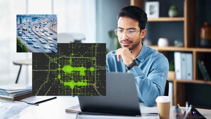 Man wearing glasses and a blue shirt sitting at his desk looking at two computer screens showing a map of a city with green dots and a photo of a busy highway