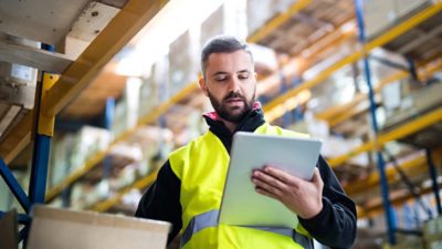 A man wearing a bright yellow vest in a warehouse looking at a tablet