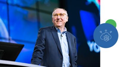 Jack Dangermond 身着深蓝色西装和浅蓝色衬衫，微笑的站在一场大型会议的讲台上，背景是显示深蓝色地图的巨大屏幕