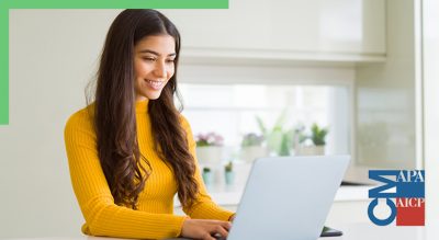 A smiling person in a bright yellow turtleneck using a laptop in a sunlit home office